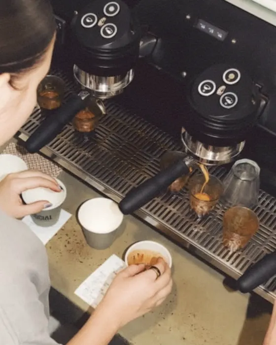 Overhead view of a cafe counter, a barista's hands wiping a cup beside the espresso machine's drip trays.