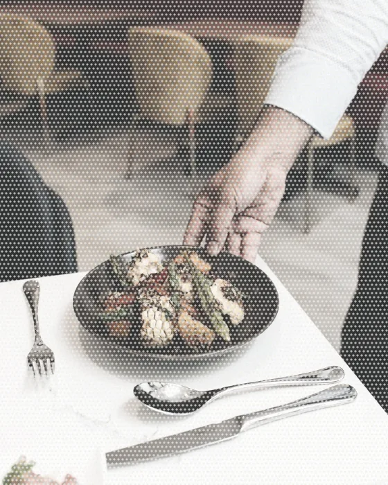 A cook's hand setting down a plate of asparagus at a laid restaurant table, cutlery beside it.