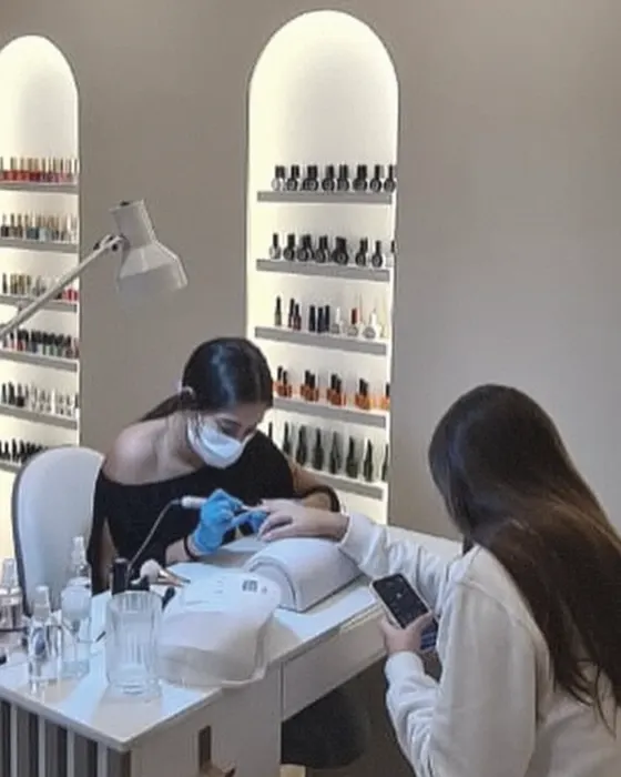 A nail technician working on a client's hand at a manicure table, shelves of polish bottles behind her.