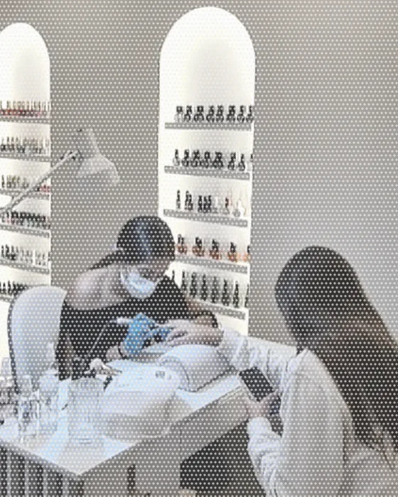 A nail technician working on a client's hand at a manicure table, shelves of polish bottles behind her.