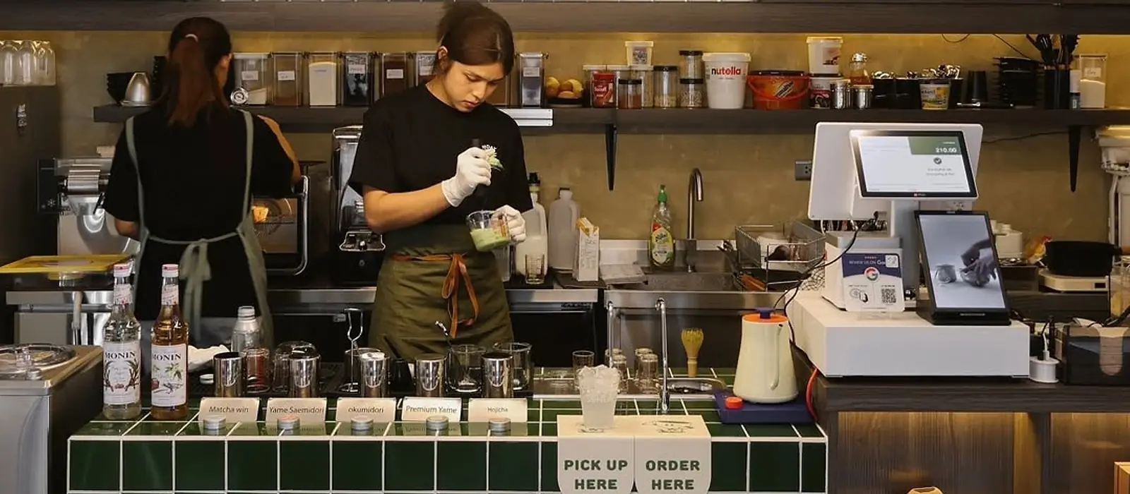 A barista in a black tee and olive apron finishing a matcha drink behind a green-tiled counter, a second staff member at the espresso machine alongside, shelves of labelled jars and a till behind them, and "Pick up here" and "Order here" signs along the counter edge.