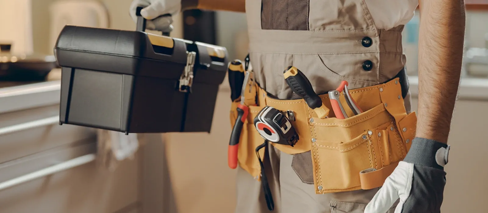 A tradesperson in overalls and a loaded tool belt carrying a toolbox through a kitchen.