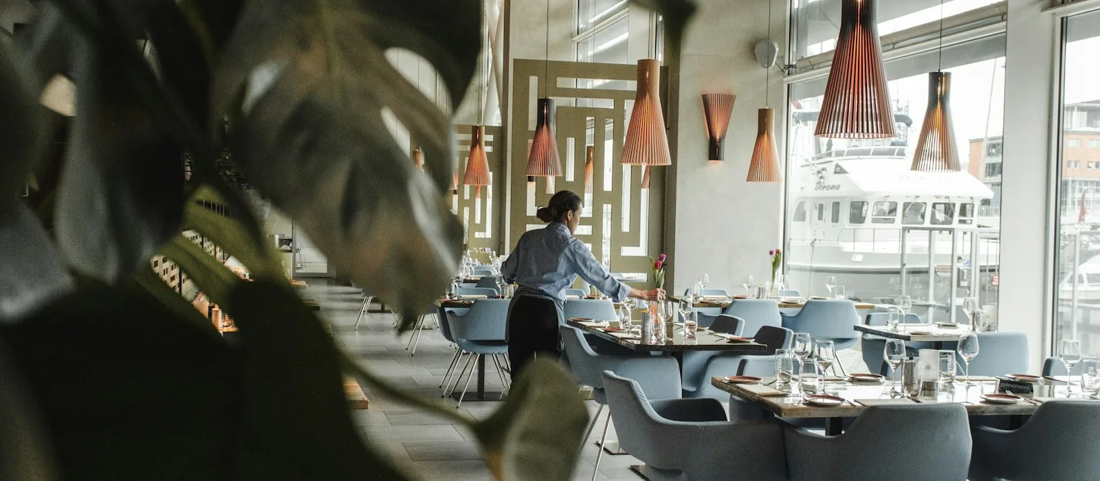 A server laying a table in a bright, high-ceilinged dining room, tables set along a wall of windows.